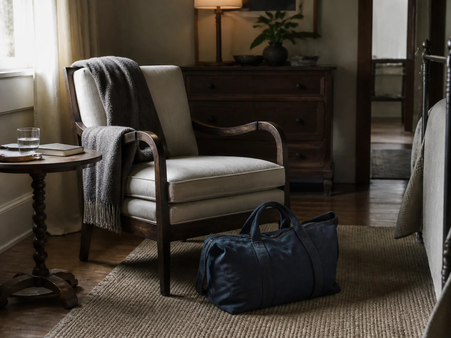 A slate-blue cloth overnight bag beside a welcoming chair, water glass and book in a warm guest room